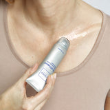 A close-up of a person's chest and neck area wearing a beige tank top, with a hand holding a silver tube of Eusthetix Elastiquy skin cream against the skin.