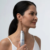 A woman with dark hair tied back in a ponytail holds a silver and white skincare bottle against her neck, shown in a close-up profile shot against a light gray background.