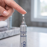 A close-up of a hand holding a clear gel drop above the applicator tip of a silver tube labeled OB MED Elastiderm.